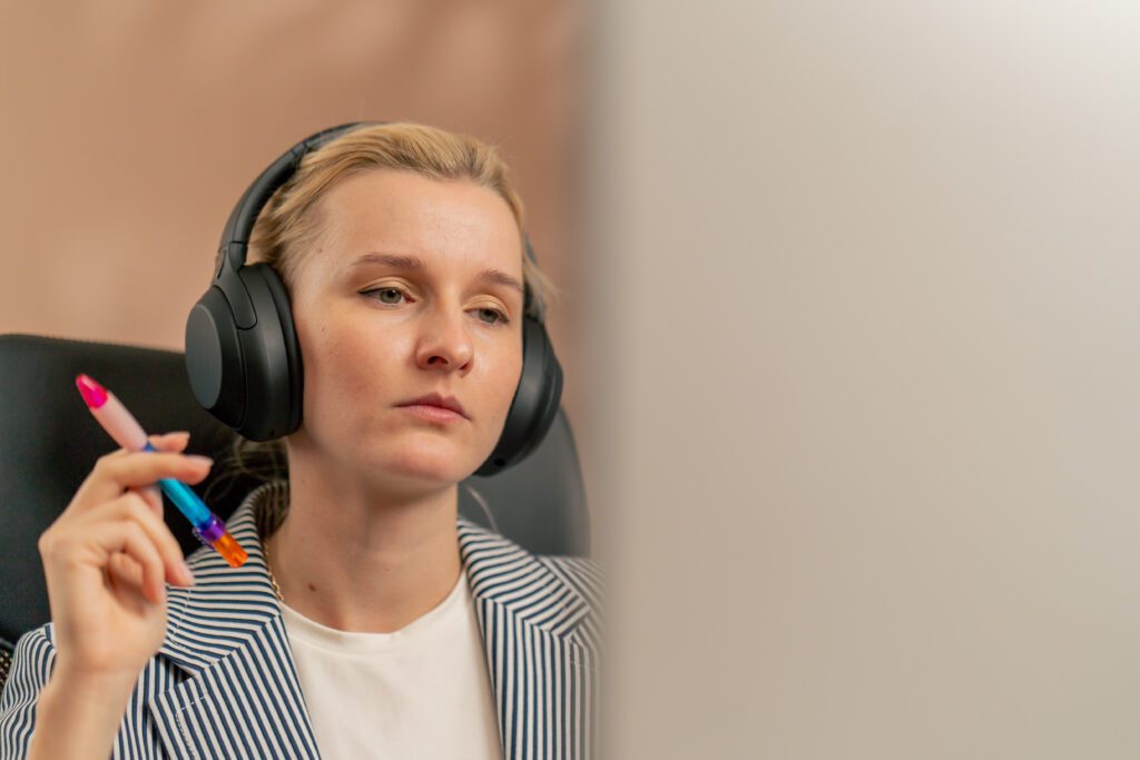 closeup in a bright office a young blonde employee works at a white monitor on a light table modern office sun rays working on a project big black headphones. Female wearing noise cancelling headphones to help decrease distractions and be more productive in the workplace.